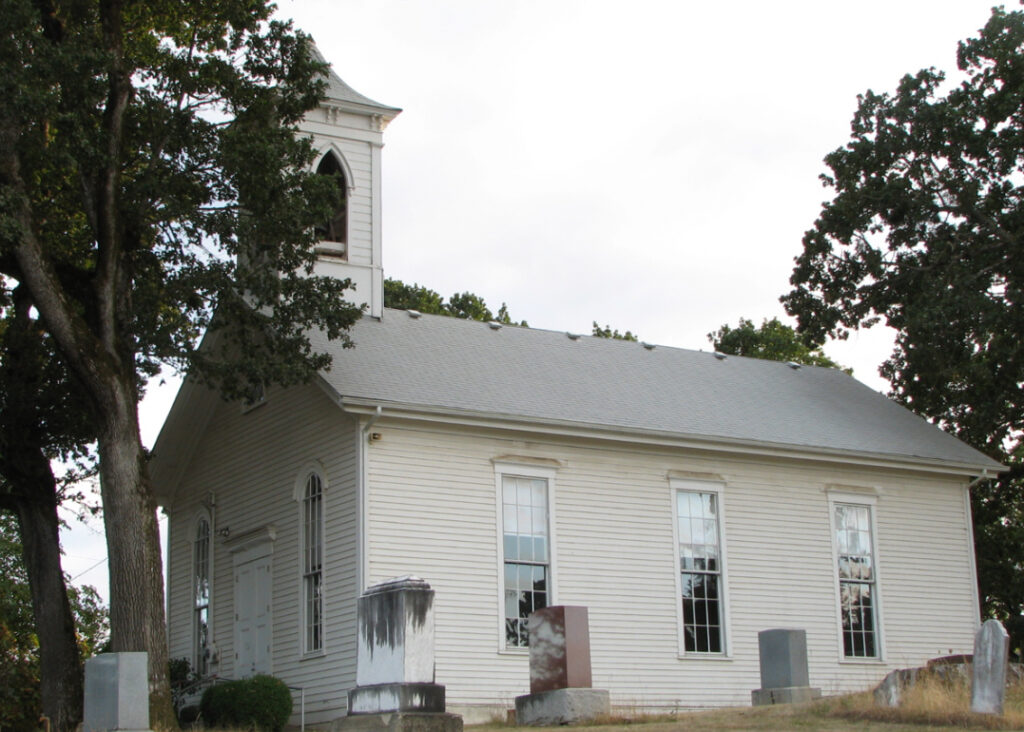 A white, traditional church building with a steeple, adjacent to a cemetery with headstones